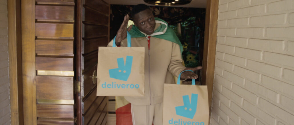 Khaby Lame portant deux sacs Deliveroo sur le pas d'une porte dans le film de la campagne Deliver Us To Fútbol pour la Coupe du Monde 2026