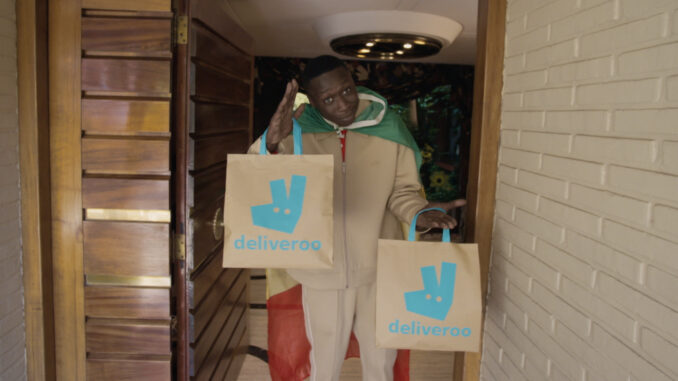 Khaby Lame portant deux sacs Deliveroo sur le pas d'une porte dans le film de la campagne Deliver Us To Fútbol pour la Coupe du Monde 2026