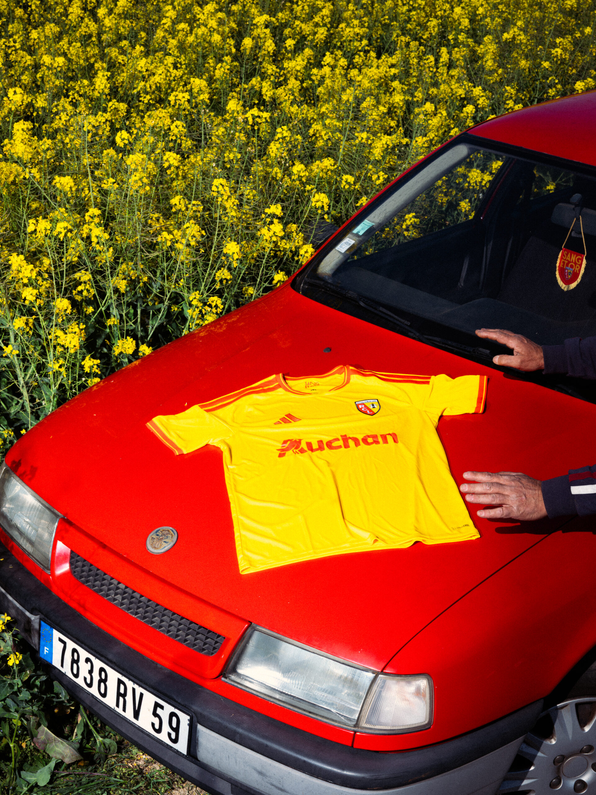 Maillot domicile adidas RC Lens 2026-2027 posé sur le capot d'une voiture rouge devant un champ de colza dans le Pas-de-Calais