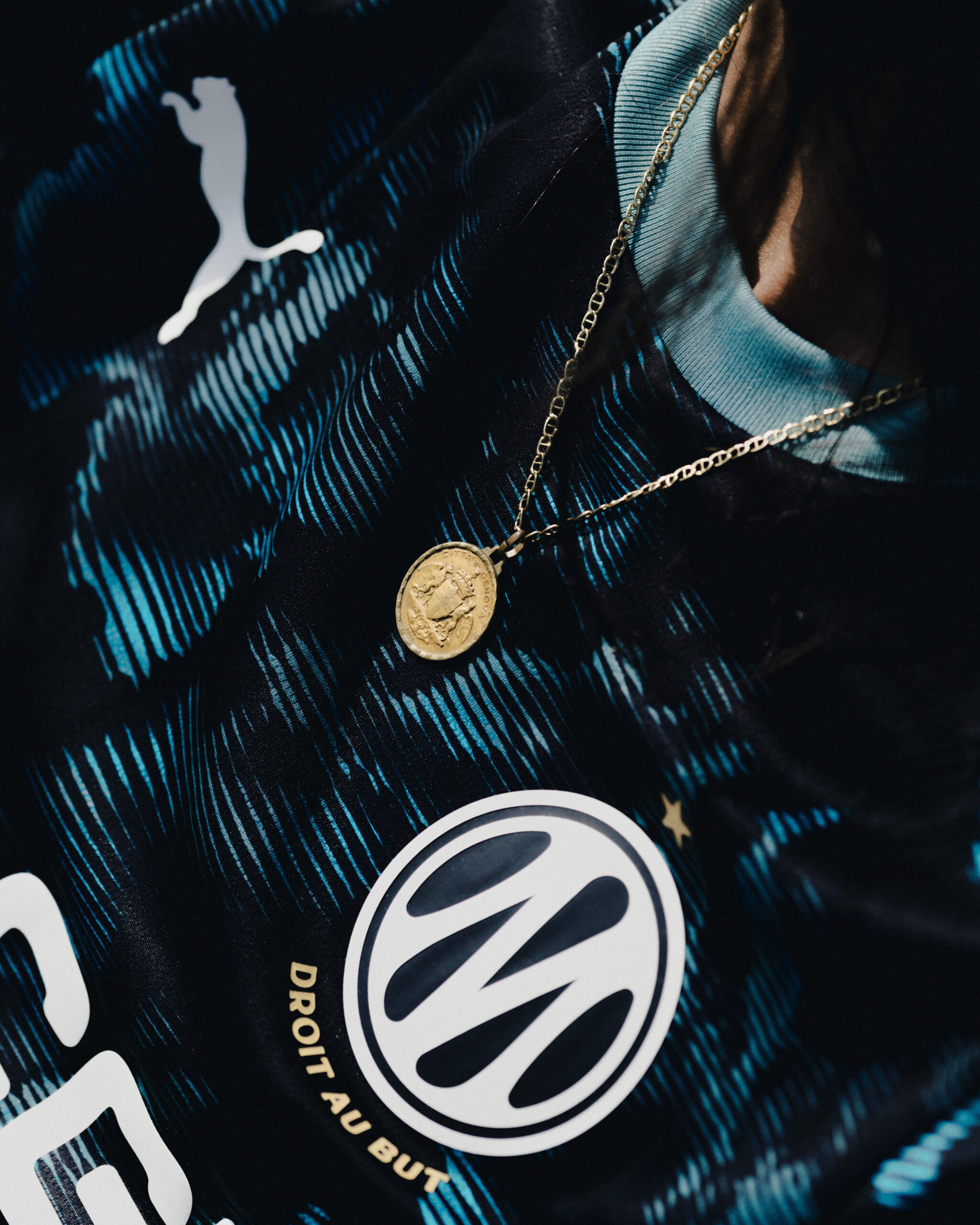 Close-up du badge OM avec médaille dorée en pendentif. Détail produit élégant. Crédit : PUMA × Olympique de Marseille