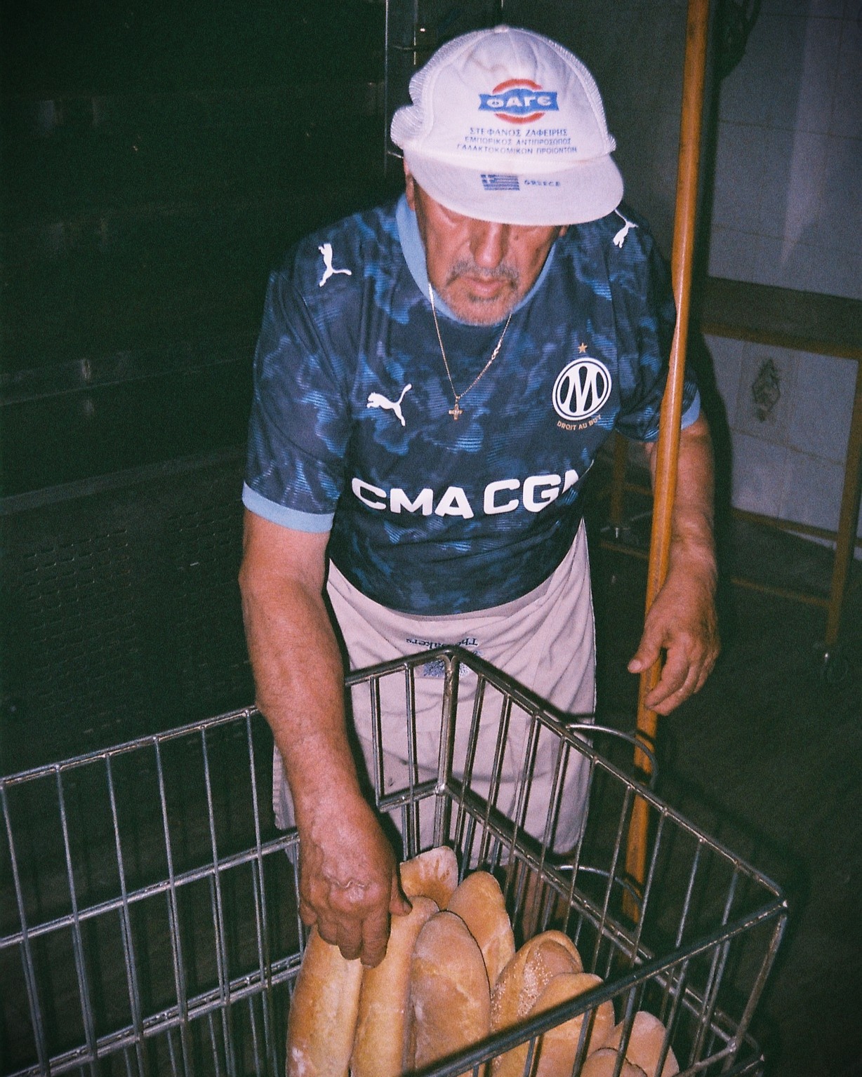 Vieil homme grec en maillot de l'OM dans sa boulangerie. Photo d'Apostolis Dimitropoulos. Crédit : PUMA × Olympique de Marseille