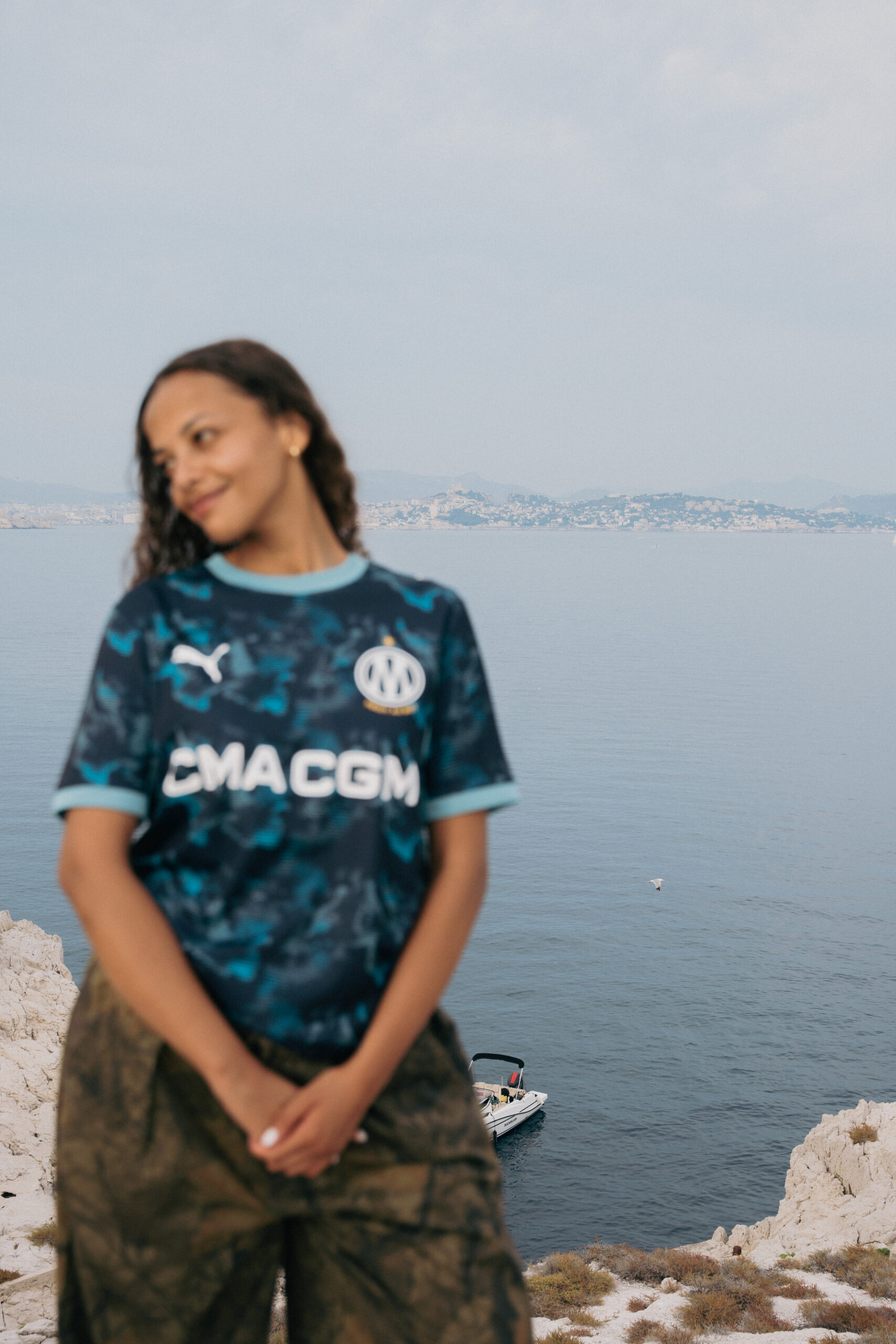 Femme portant le maillot devant les calanques de Marseille. Fond de mer et de falaises reconnaissables. Photo prise depuis les hauteurs. Crédit : PUMA × Olympique de Marseille