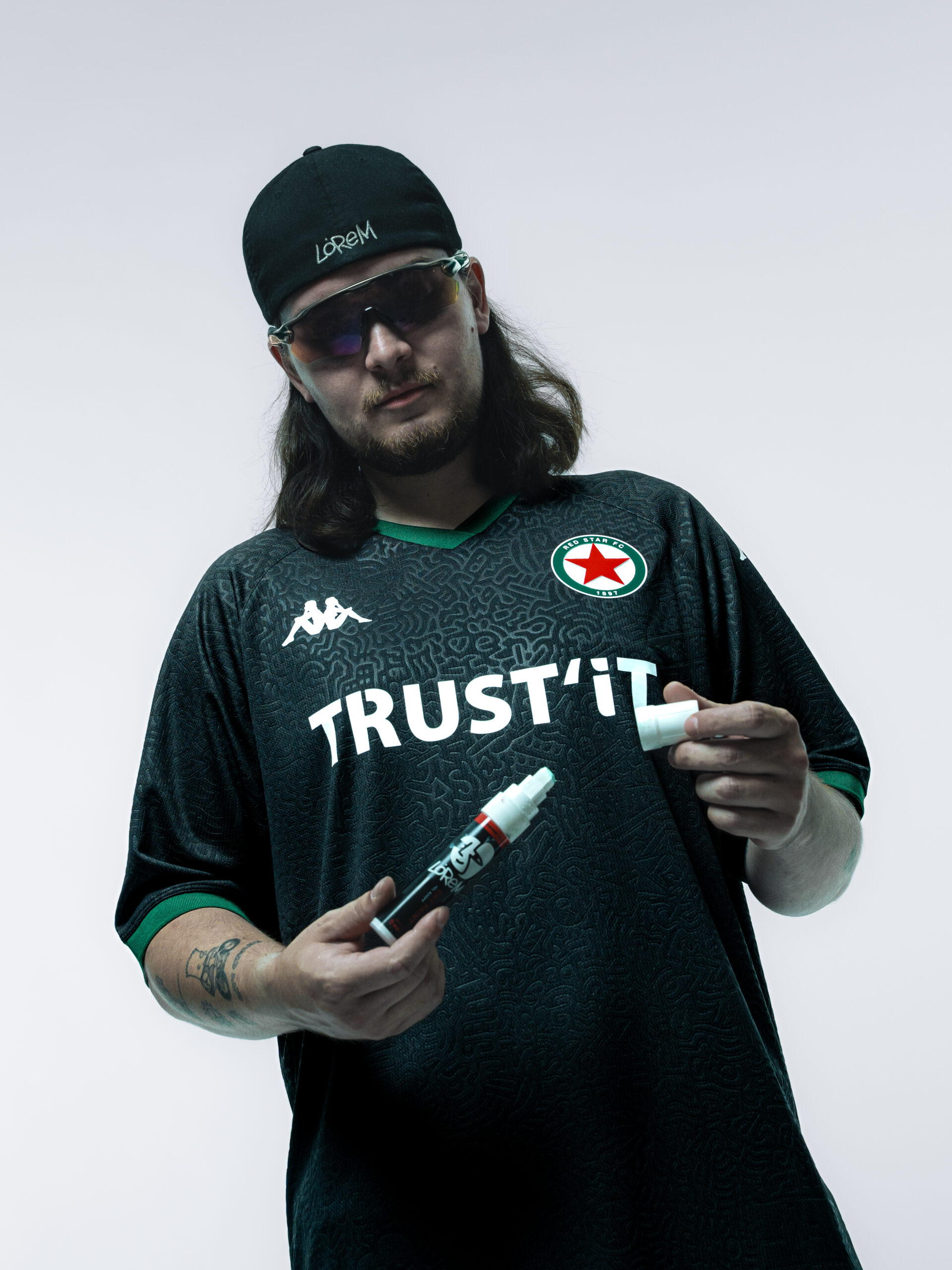 Le street-artiste Lorem tient deux marqueurs de sa marque, outils de sa pratique du doodle art, en portant le maillot Le Vert & Noir du Red Star FC