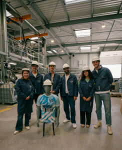 Six personnes en veste Recyc'Elit et casque blanc posent en usine autour d'un mannequin portant le Maillot Infini bleu de NOLT